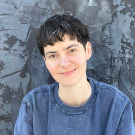 Person with short dark hair smiling in a blue shirt against a gray textured wall