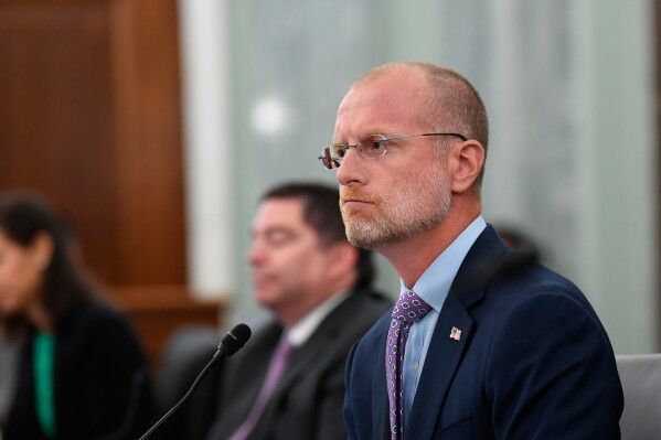 Man with glasses and a beard in a suit at a hearing, looking to the side.