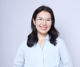 Smiling woman with glasses and black hair wearing a light blue blouse against a pale background