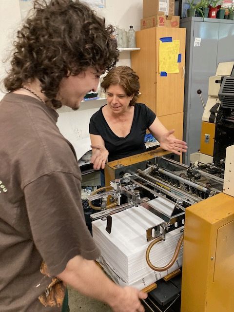 Man and woman look at a printing press. Man holds stack of paper, smiling. Woman gestures with hands, looking on.