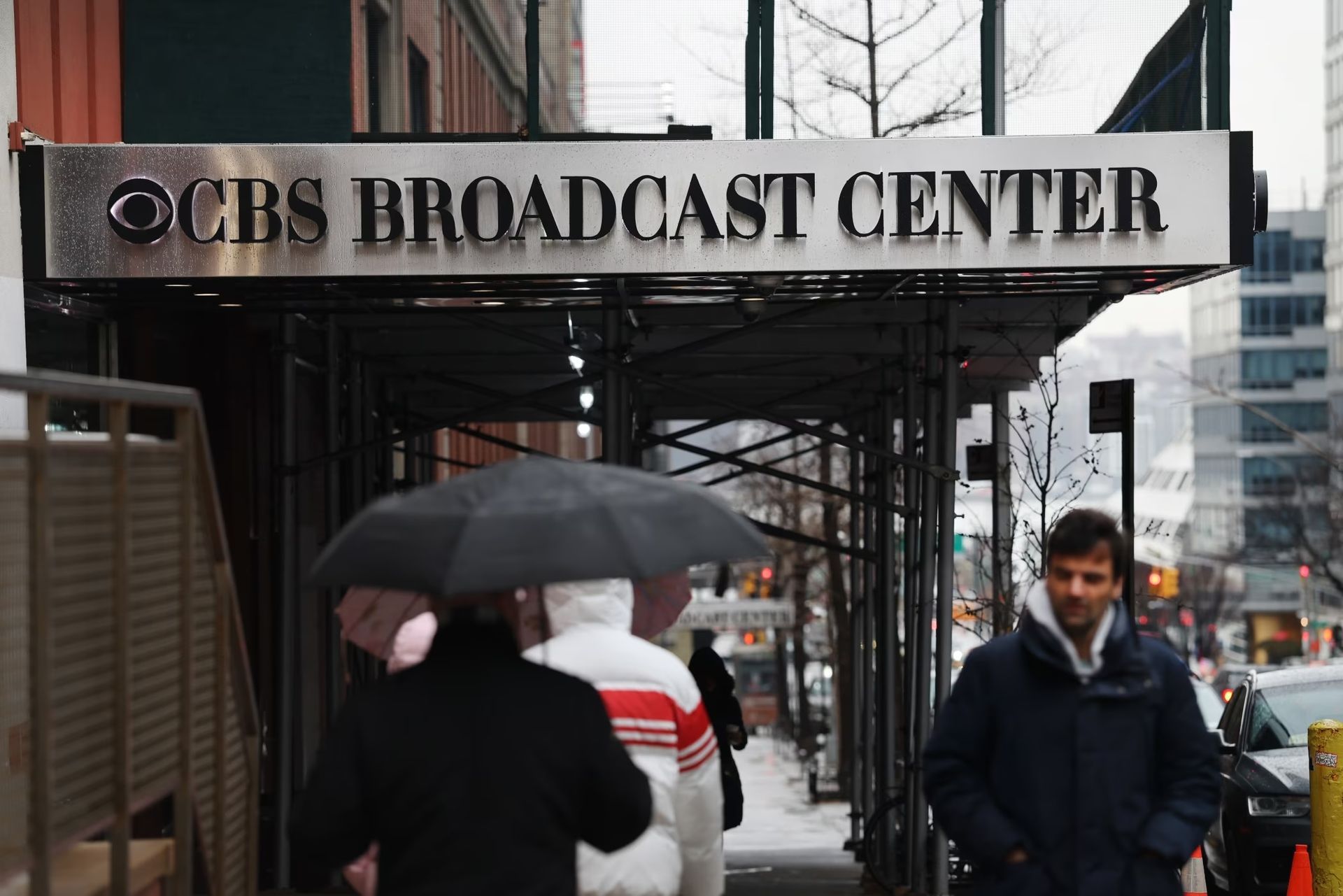 People walk past the CBS Broadcast Center entrance under scaffolding on a winter day.