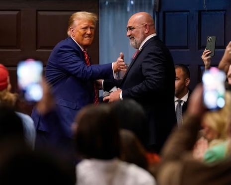 Donald Trump shaking hands with a man in a suit, both smiling, surrounded by a crowd.