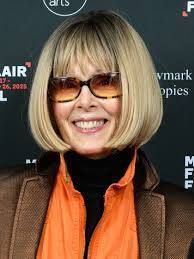 Smiling person with blonde bob haircut, sunglasses, orange scarf, and brown jacket at an event backdrop