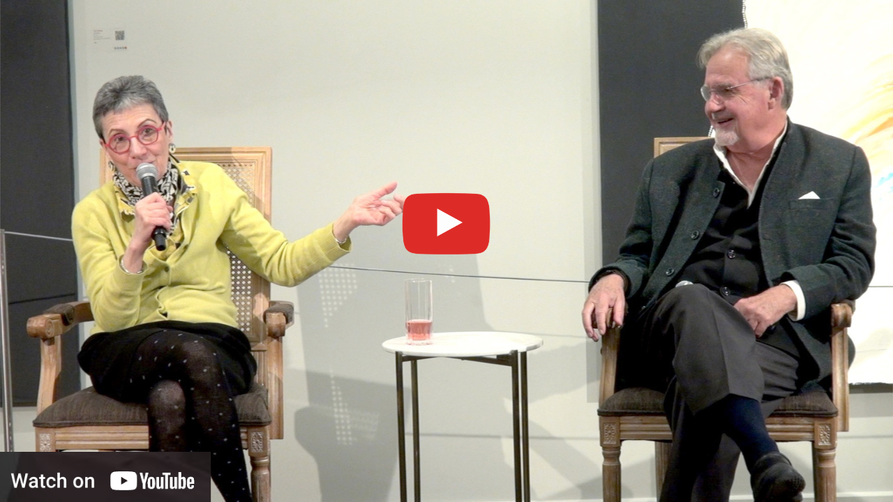 Two people sit in chairs on a stage during a presentation, with a speaker holding a microphone and gesturing to the side.
