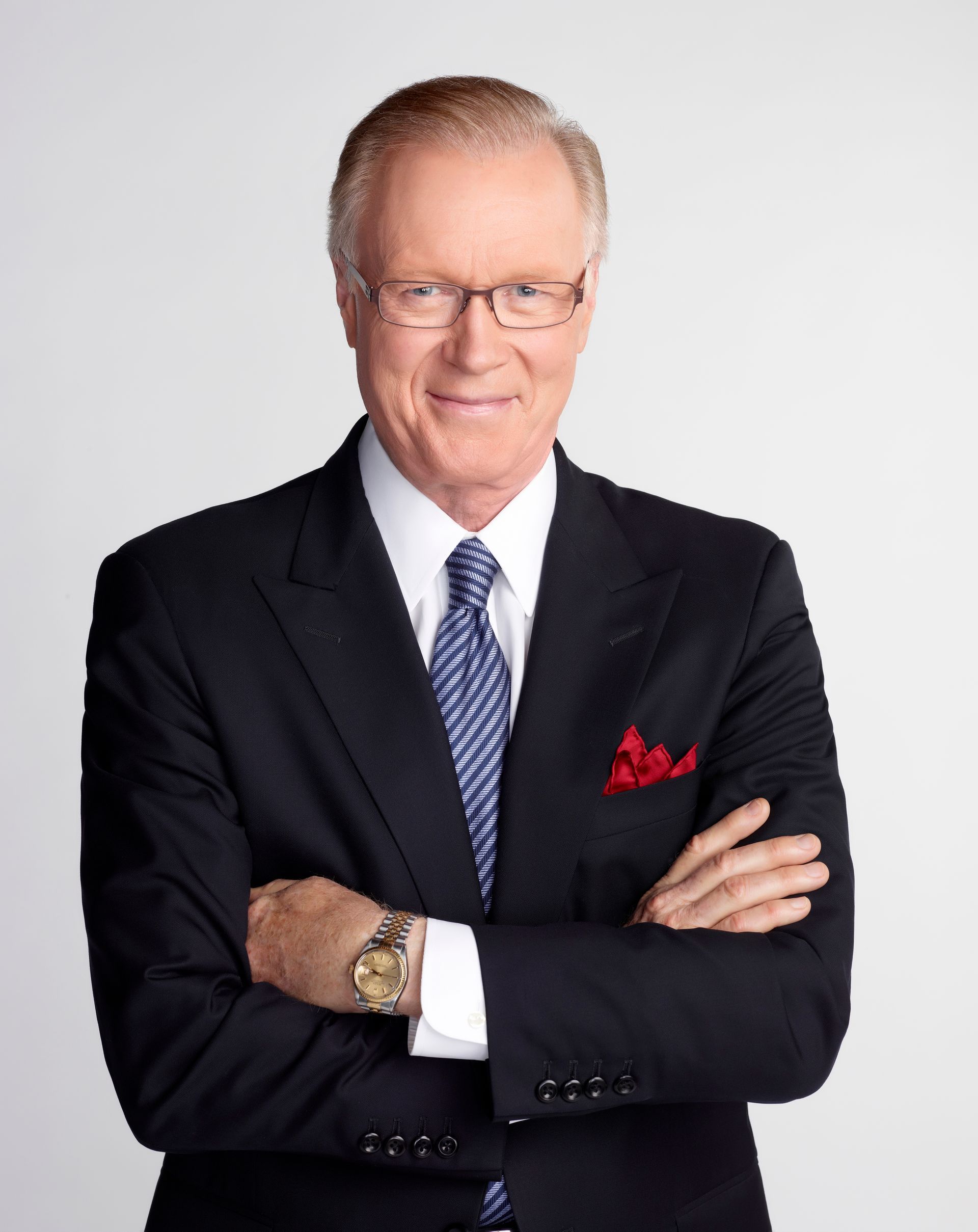 A person with gray hair wearing a dark blazer and white shirt stands with hands in their pockets against a gray backdrop.