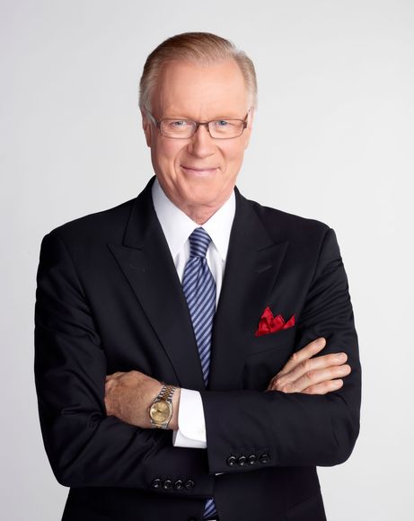 A person with gray hair wearing a dark blazer and white shirt stands with hands in their pockets against a gray backdrop.