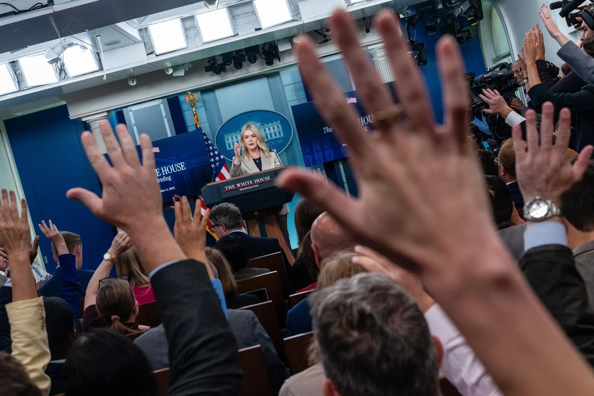 White House press briefing: Woman speaking at podium, American flag in background, hands raised.