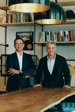 Two people standing behind a wooden table in a modern office with shelves and hanging lamps.