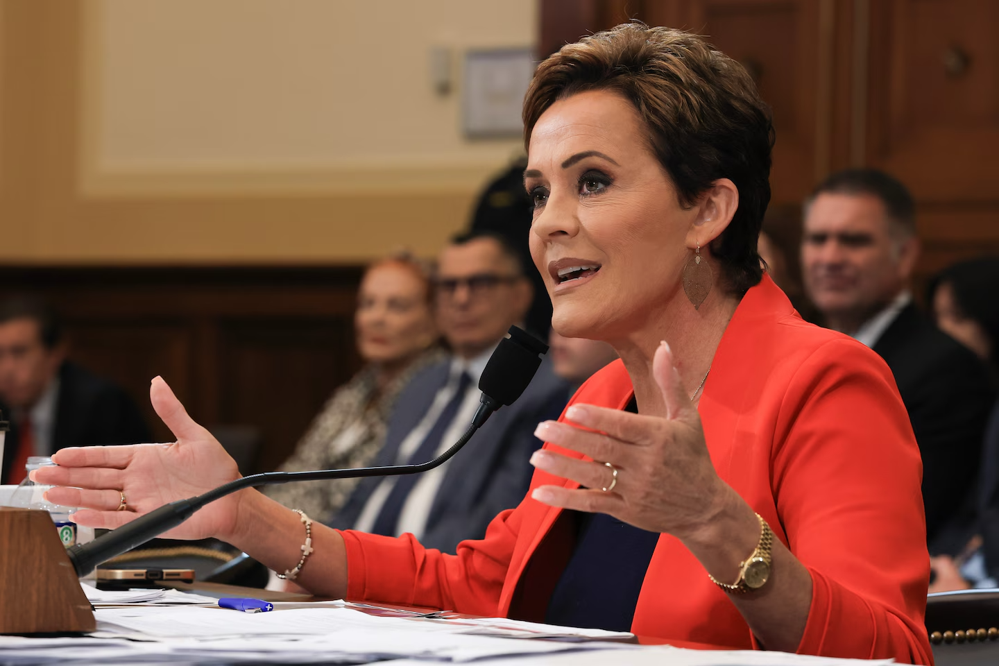 Kari Lake at a hearing, wearing a red blazer, gesturing with hands while speaking into a microphone.