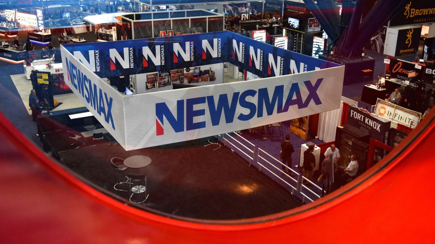 Newsmax banner hangs over a booth, blue letters on white. Inside, people and merchandise.