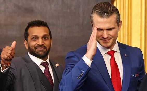 Two men in suits pose indoors, one giving a thumbs-up and the other a salute.