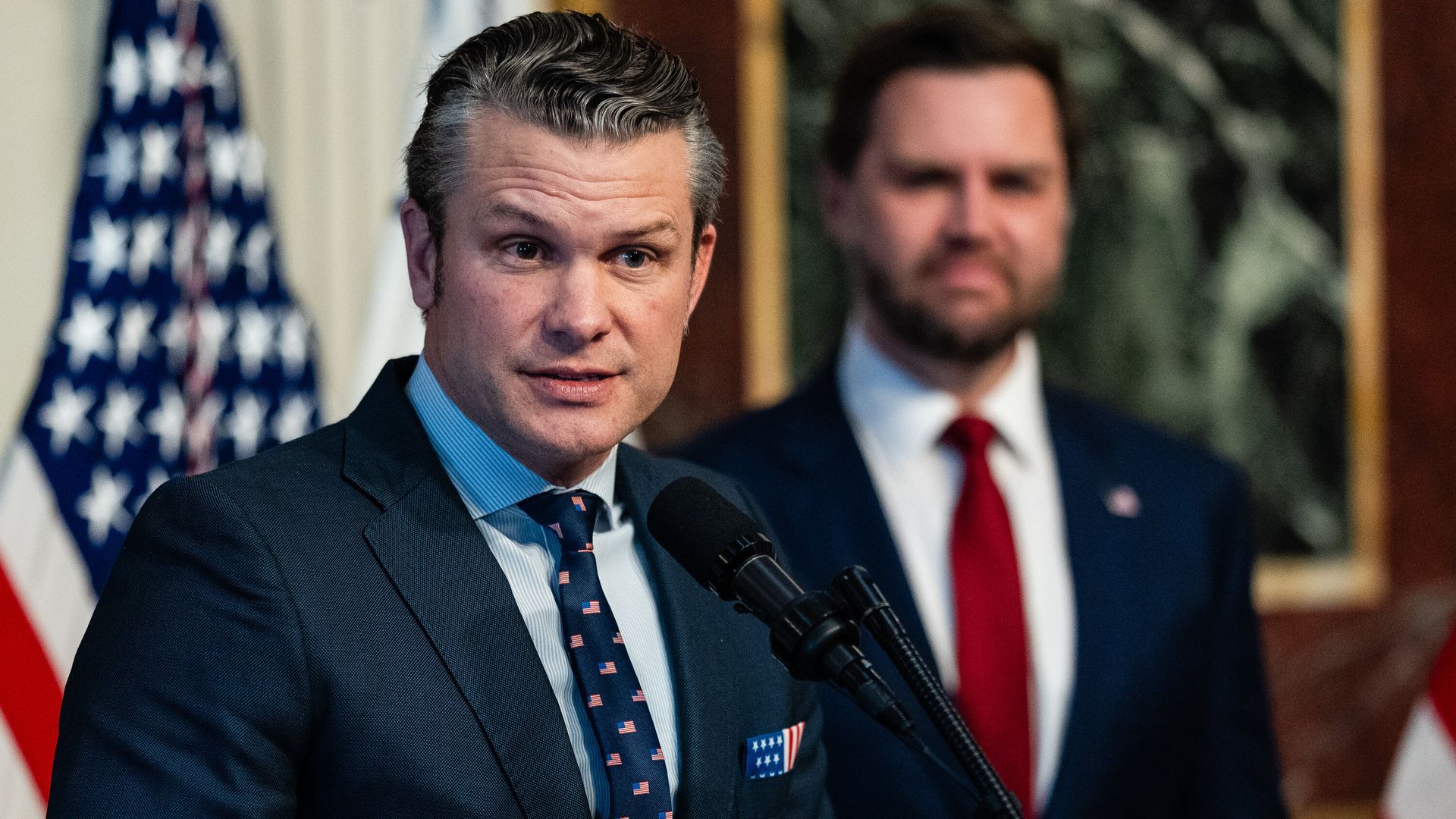 Man speaking into microphone with another man standing behind in front of US flag.