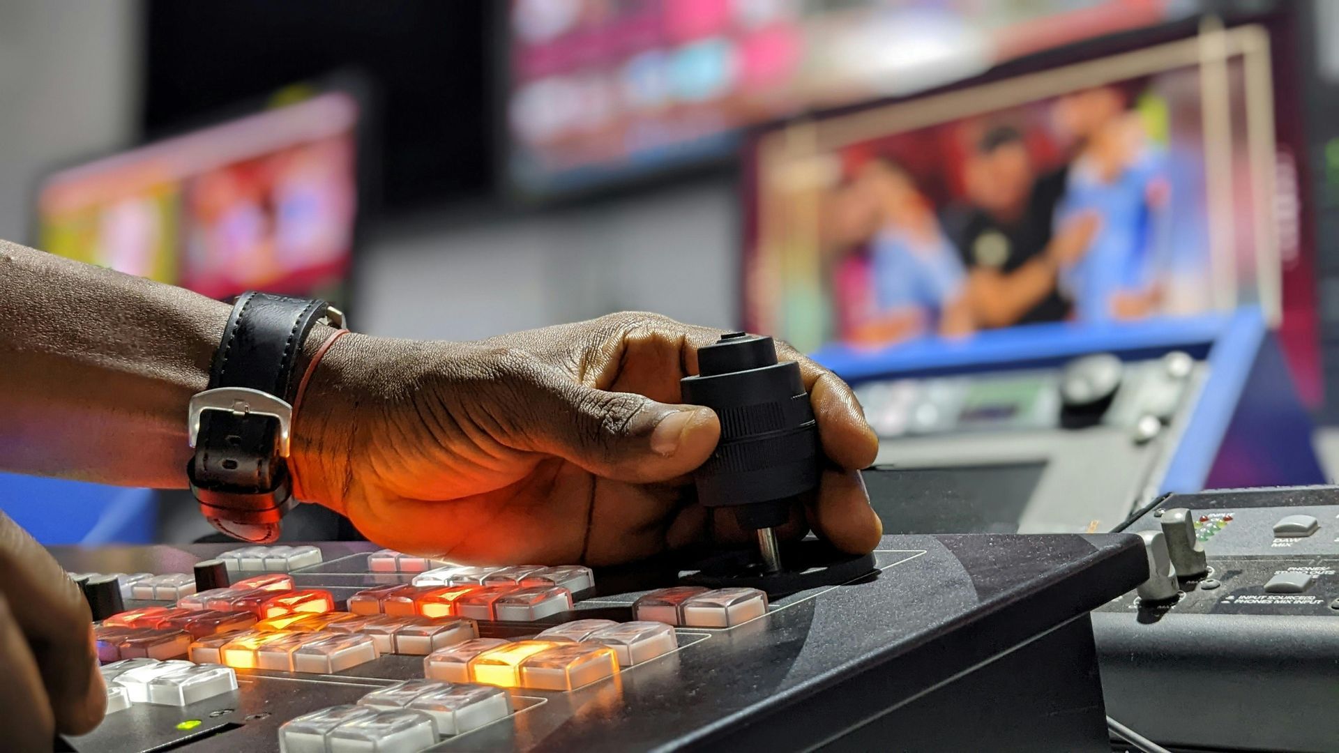 A person's hand operates a control panel with lit buttons; monitors with a live feed are in the background.
