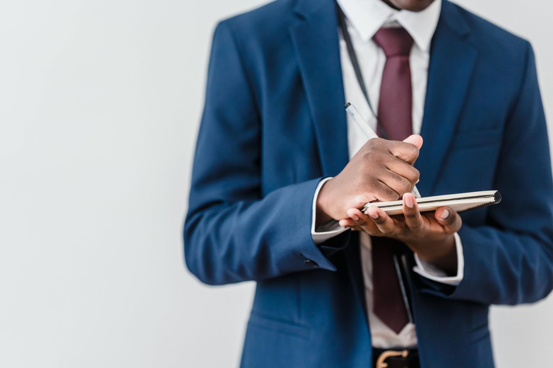 Man in blue suit writes in a notebook, wearing a red tie and lanyard, against a plain background.