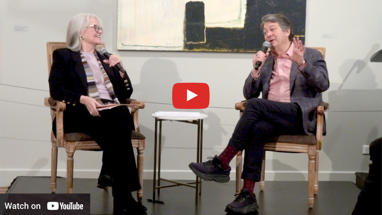 Two people seated, conversing. One holds a microphone, the other smiles. They are in a gallery setting with abstract art.