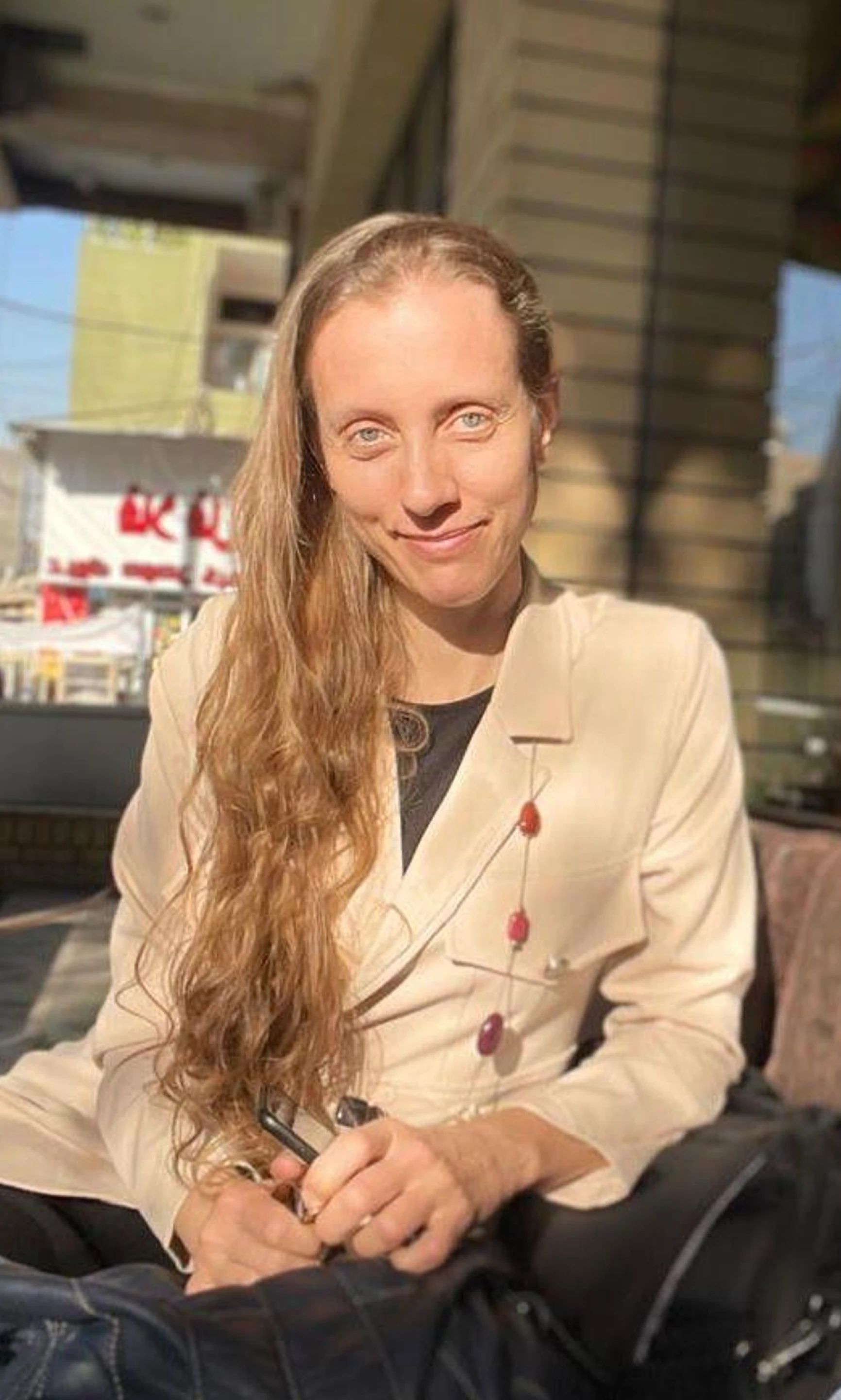 A person with long blonde hair, wearing a beige blazer, smiling while sitting outdoors in natural sunlight.