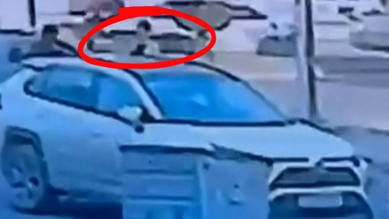 A low-quality surveillance image showing a person standing in the open sunroof of a white SUV.