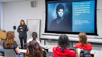 Woman presenting in a classroom with a projection screen. Students listen. Screen shows a woman and text.