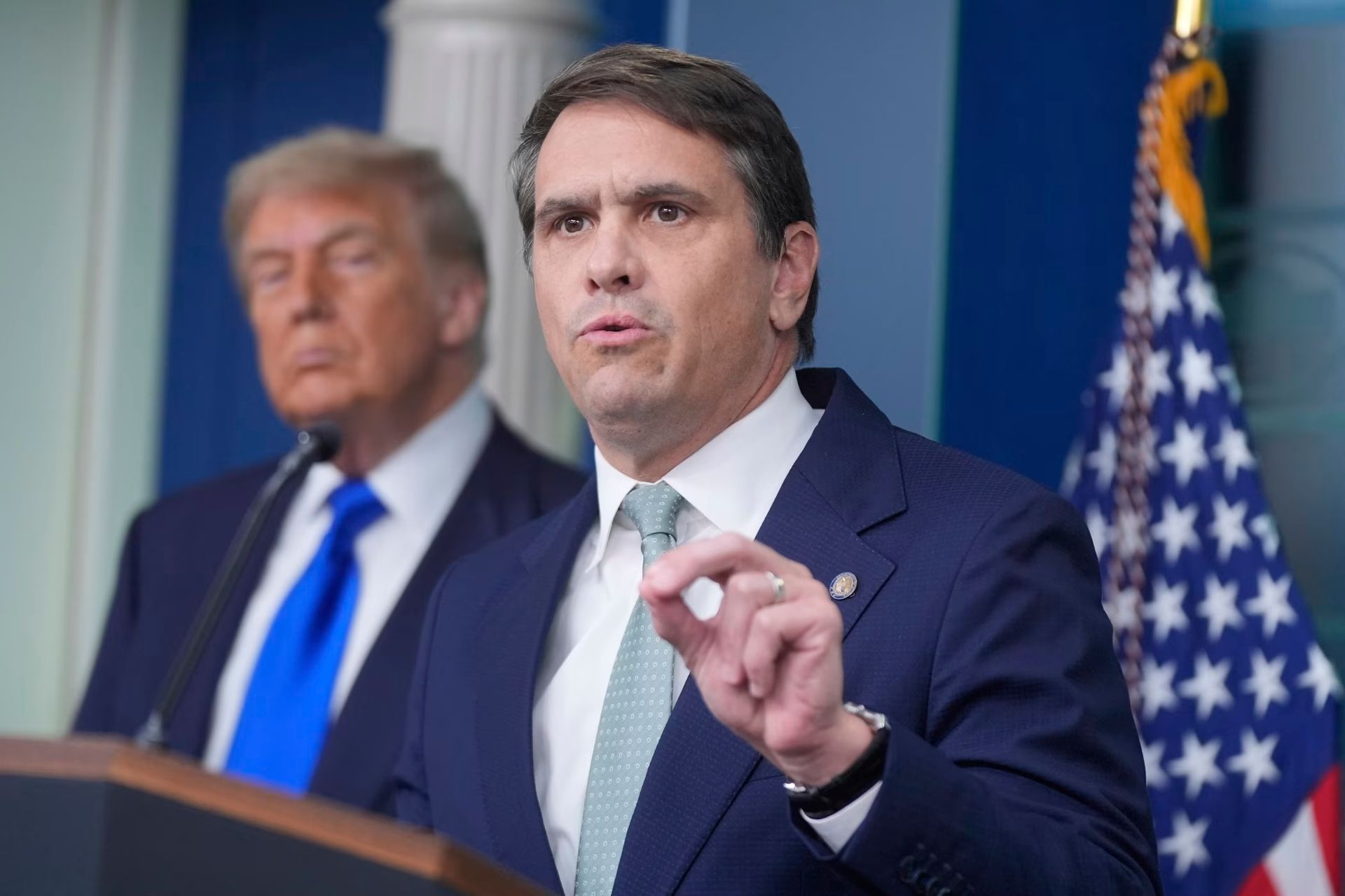 Man in navy suit speaks at podium with Donald Trump blurred in background, U.S. flag behind them.