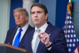 Man in navy suit speaks at podium with Donald Trump blurred in background, U.S. flag behind them.