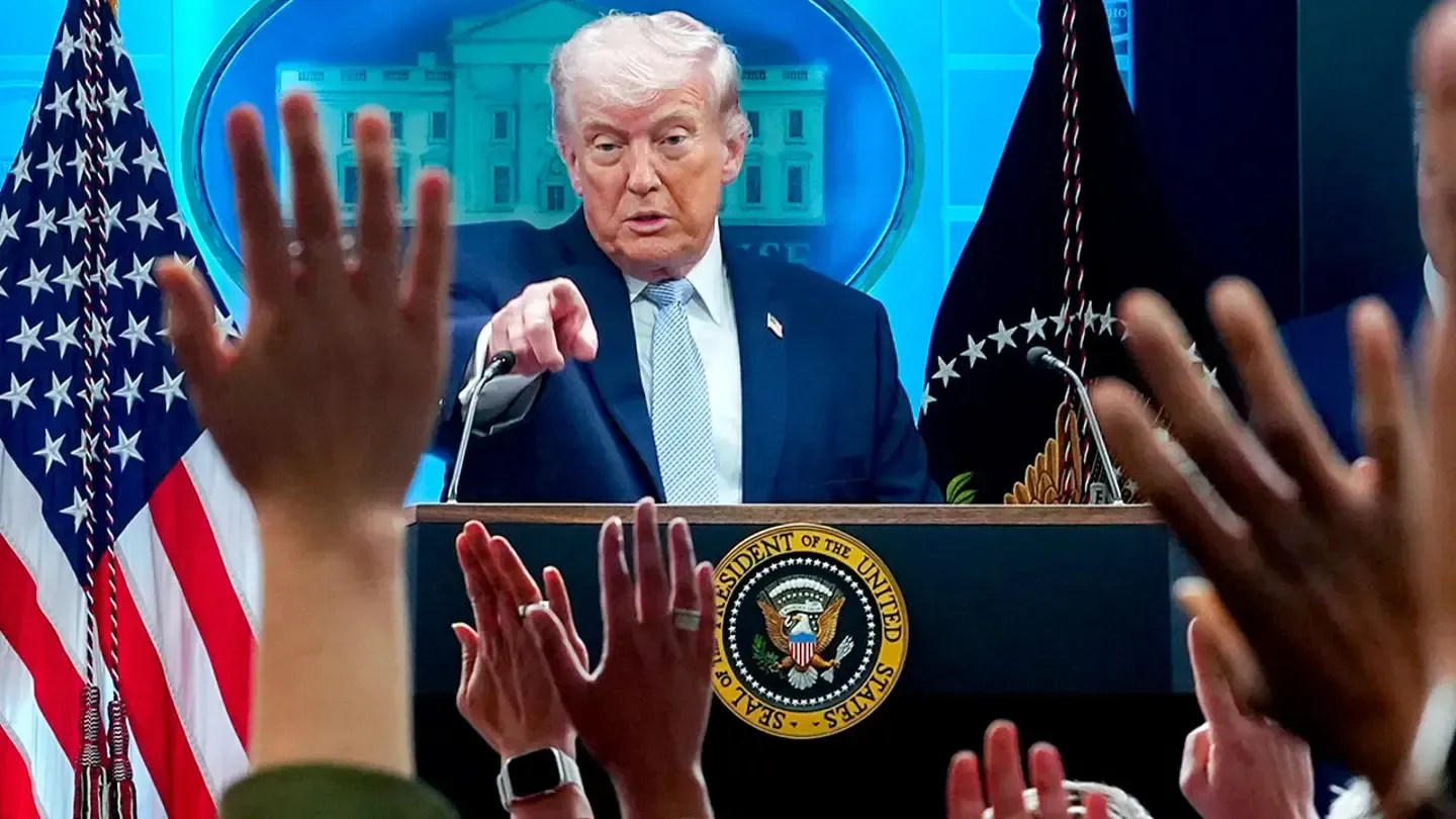 Donald Trump speaking at a podium with U.S. flags, with raised audience hands in the foreground