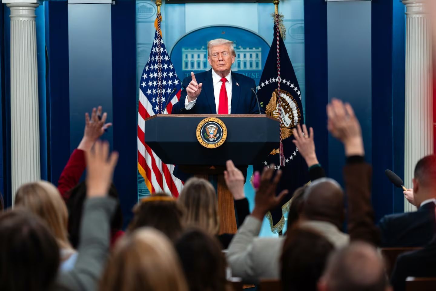 Donald Trump at a White House podium, gesturing, answering questions during a press conference. Journalists raise hands.
