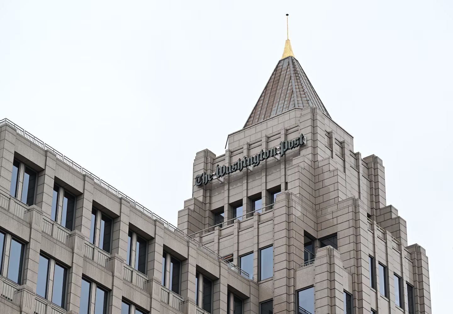 The exterior entrance of The Washington Post headquarters building, framed by lush green trees under a bright blue sky.