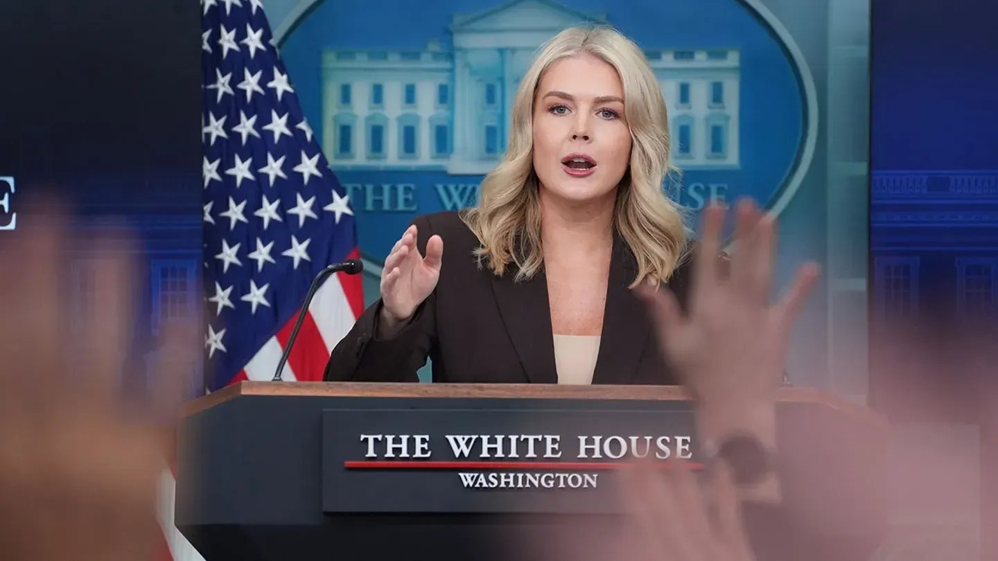 White House press briefing: A woman speaks at a podium with raised hands in the foreground. American flag and White House backdrop.