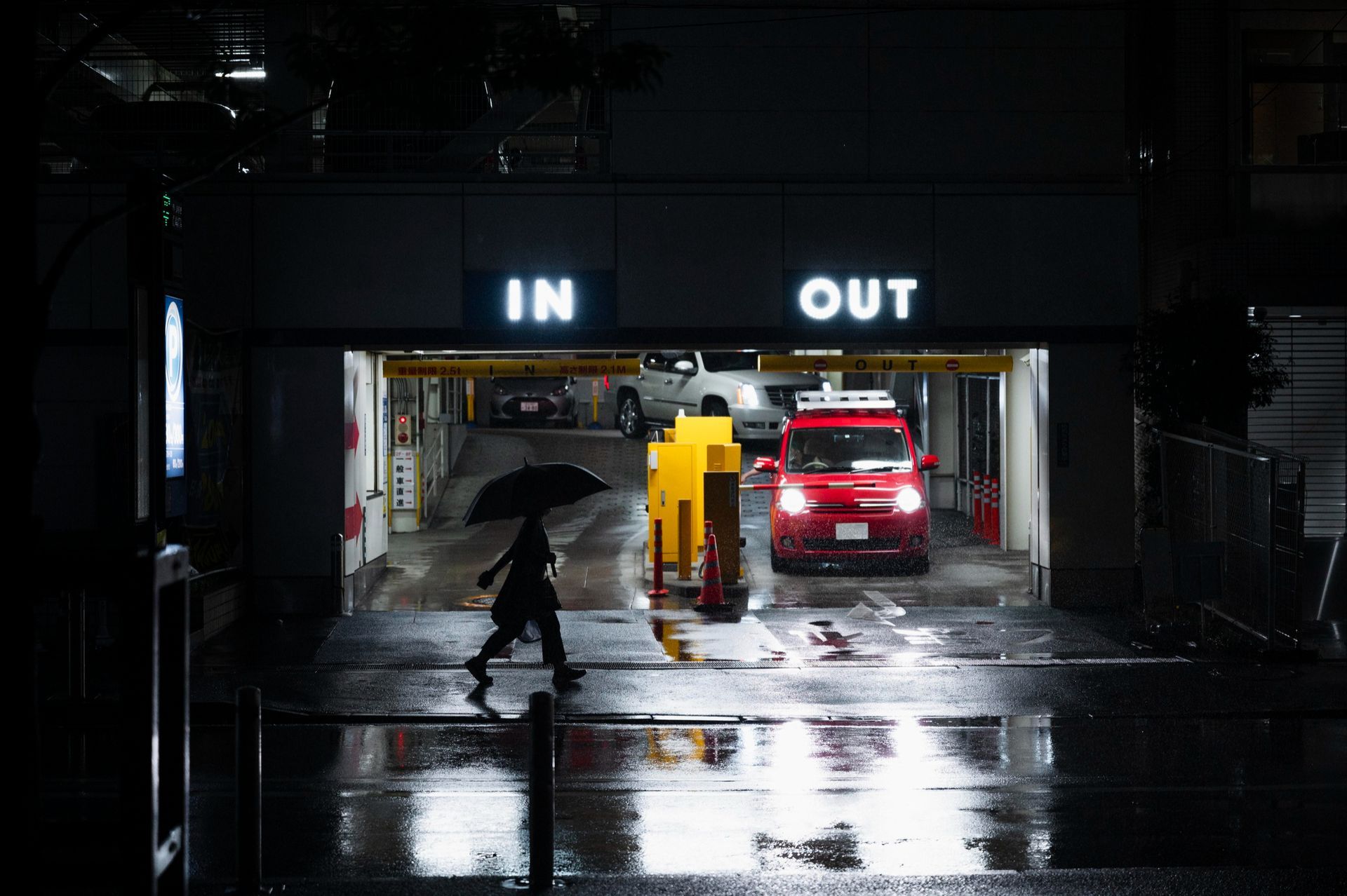 Person with umbrella walks across a wet street toward a parking garage entrance; red car exits; 