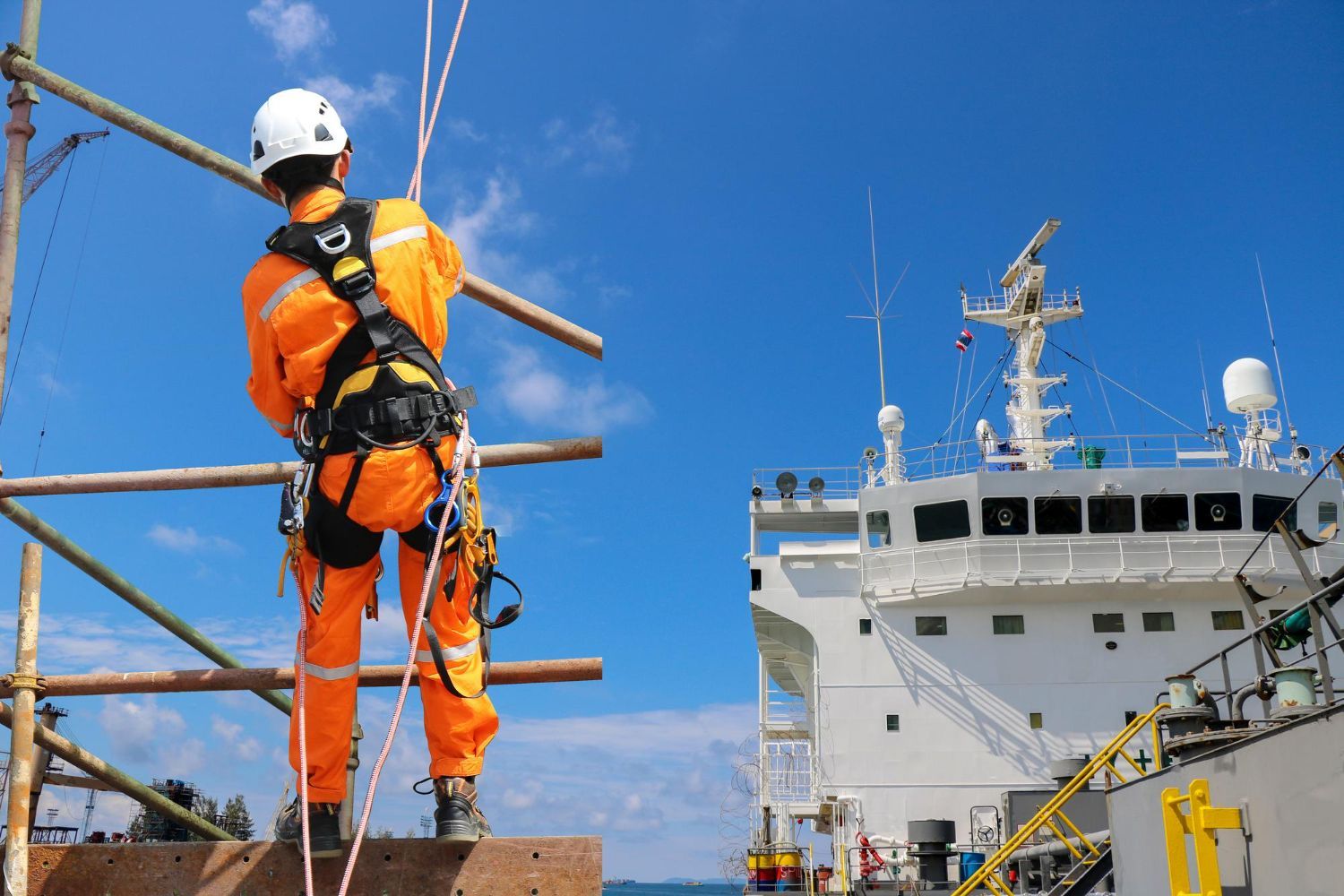 Person in orange safety suit, secured by ropes, on scaffolding near a ship under a blue sky. 