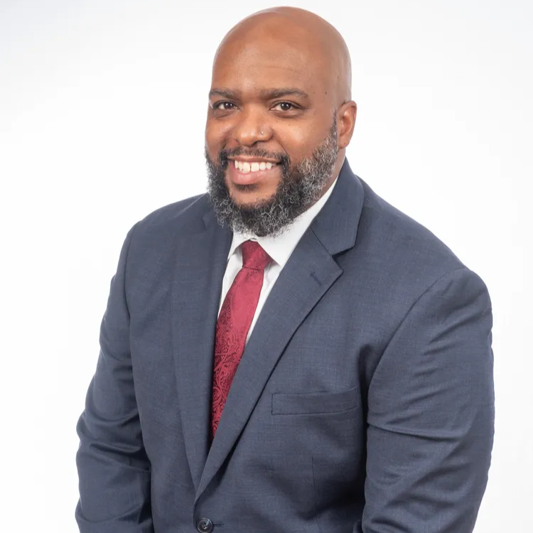 Man with dark skin, wearing a blue suit and red tie, smiling. White background.
