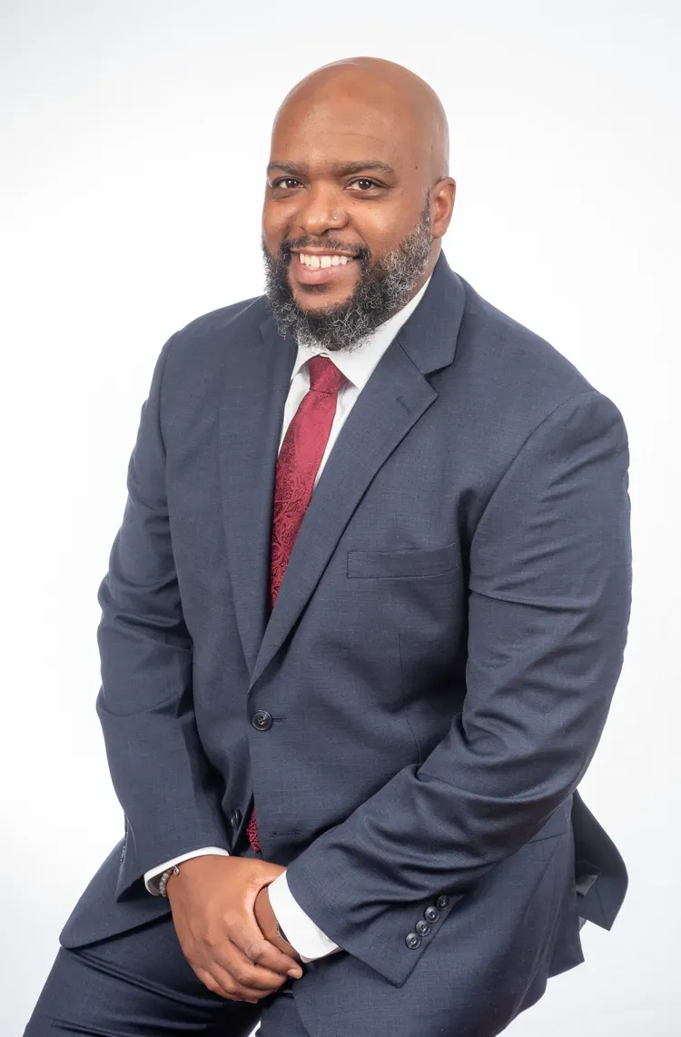 Man in navy suit, red tie, smiling; seated against a white backdrop.