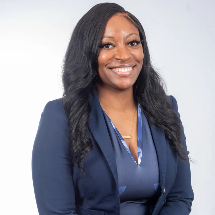 Woman with long dark hair, wearing a navy blue blazer and dress, smiles in front of a white background.
