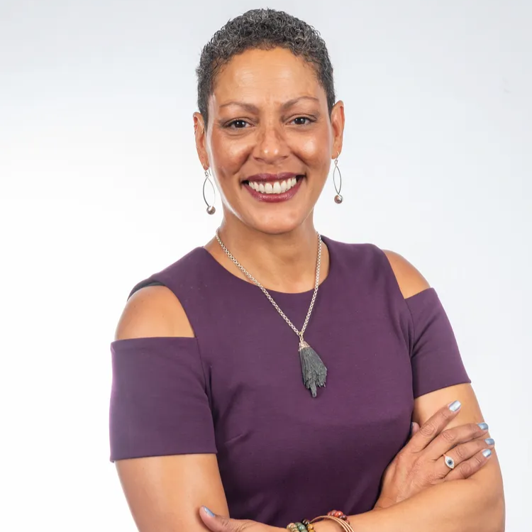 Woman in a purple dress with a necklace, smiling at the camera, arms crossed.