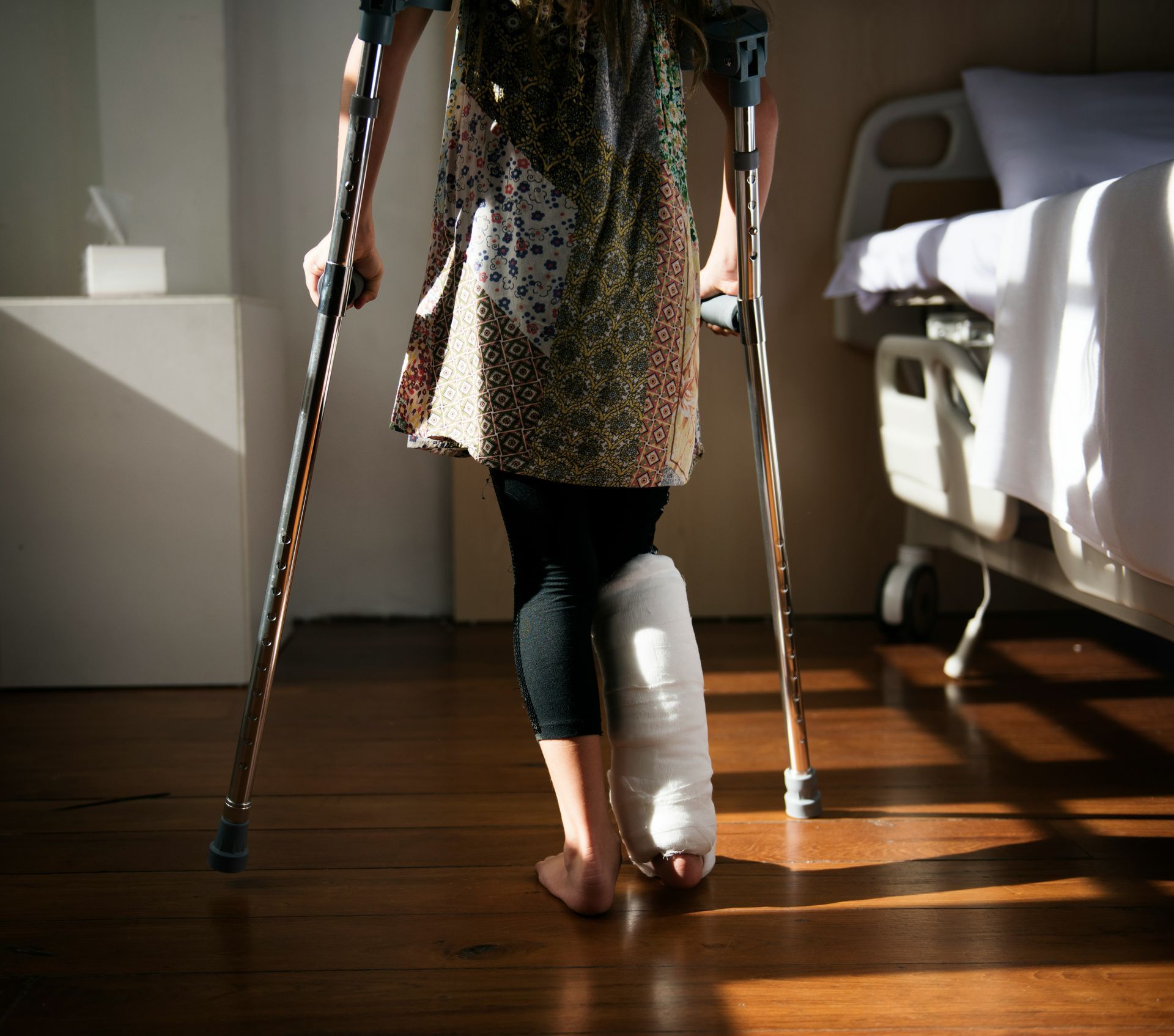 Person on crutches with leg in cast, walking on wooden floor in a hospital room.