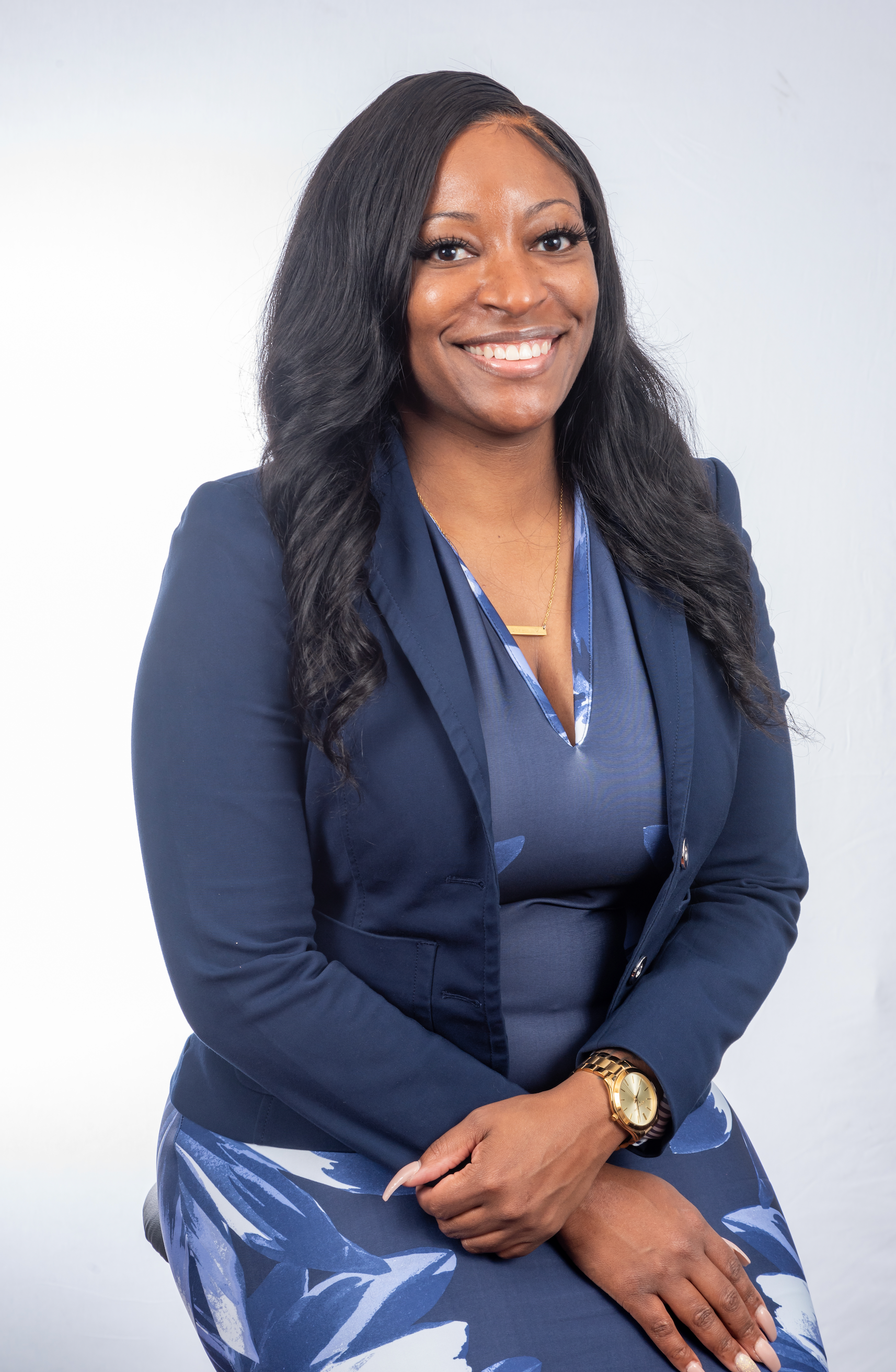 Woman in a navy suit smiles at the camera, gold watch on wrist, white background.