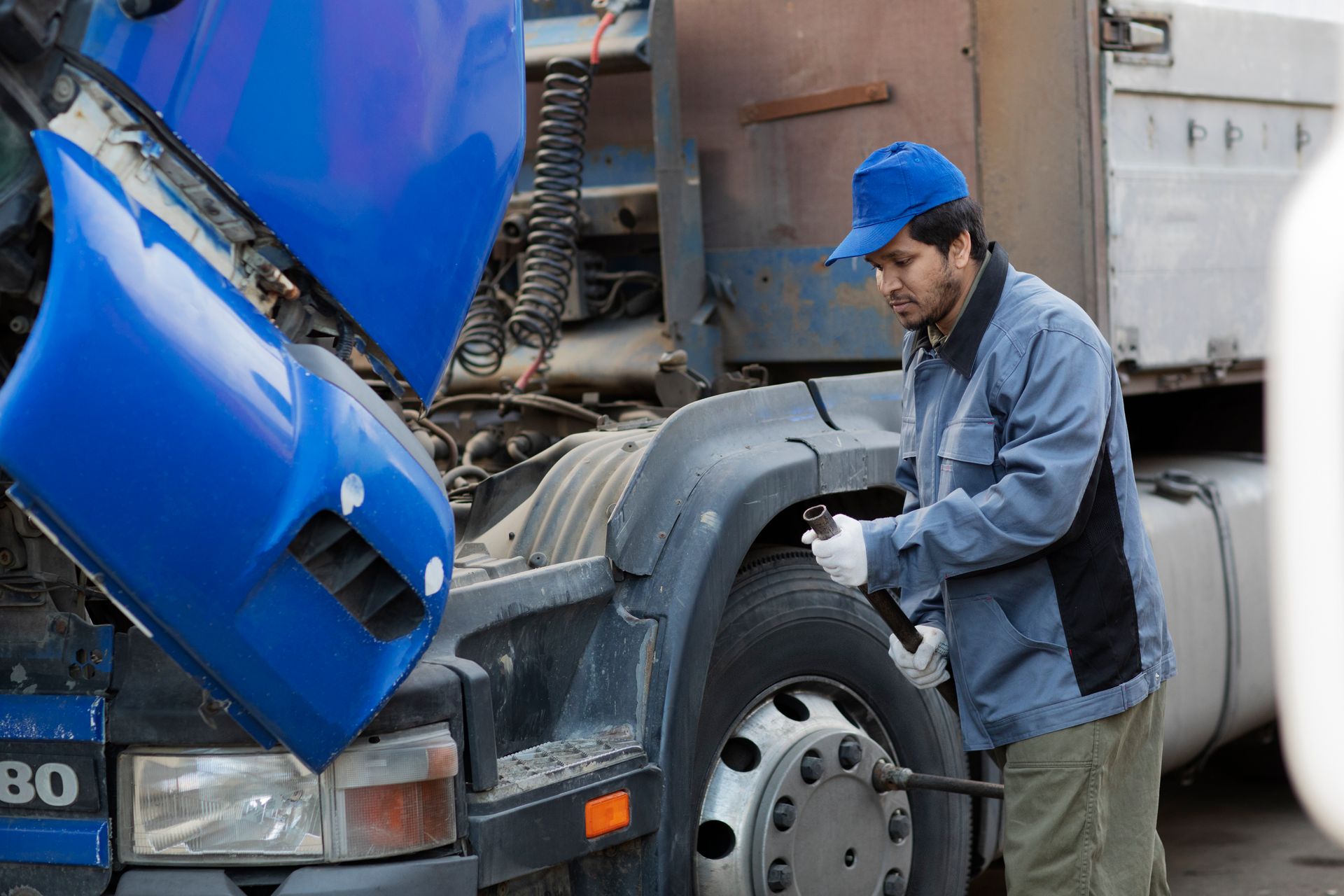 Mechanic in blue hat and jacket working on a truck tire, hood open, outdoors.