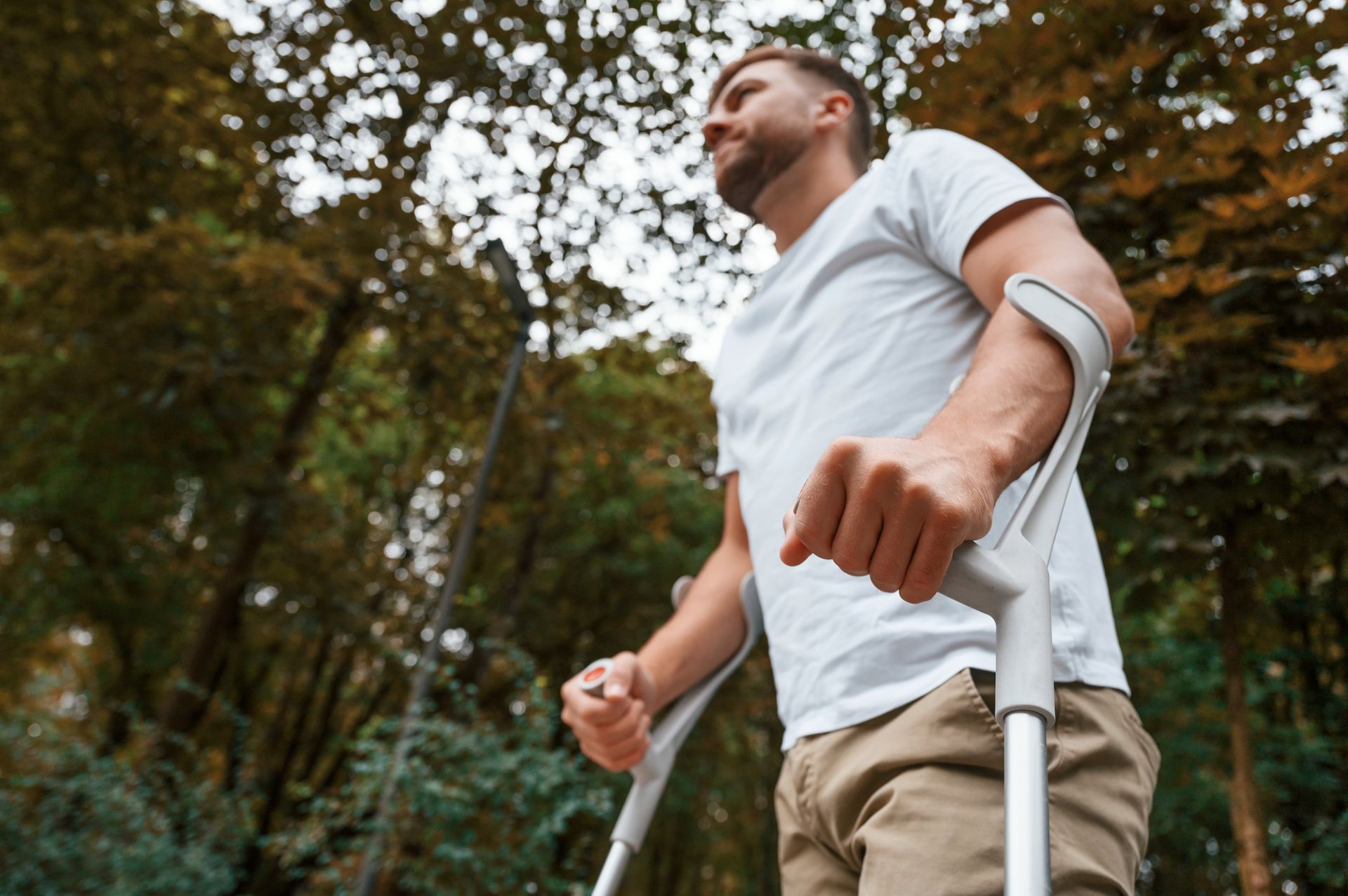 Man with crutches walking outdoors, looking up. Trees in the background.