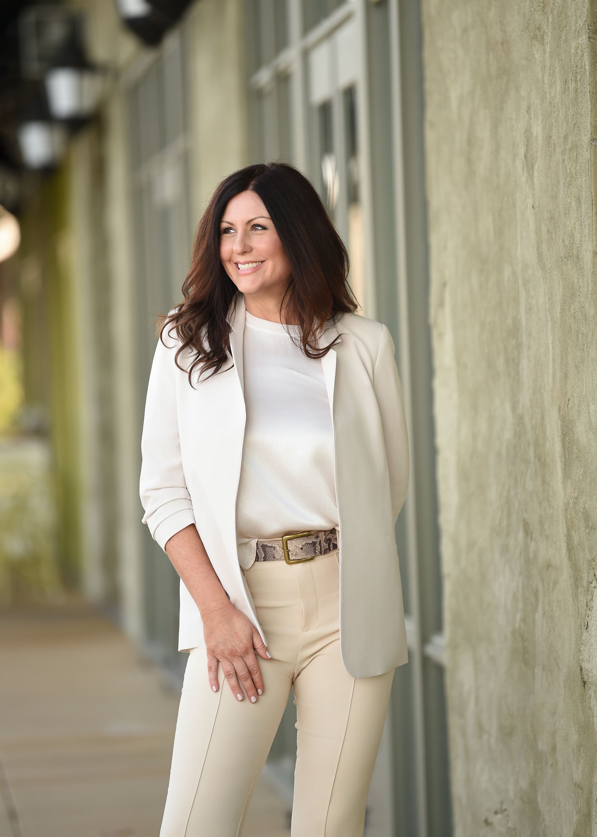 A woman in a white jacket and white pants is standing in front of a building.