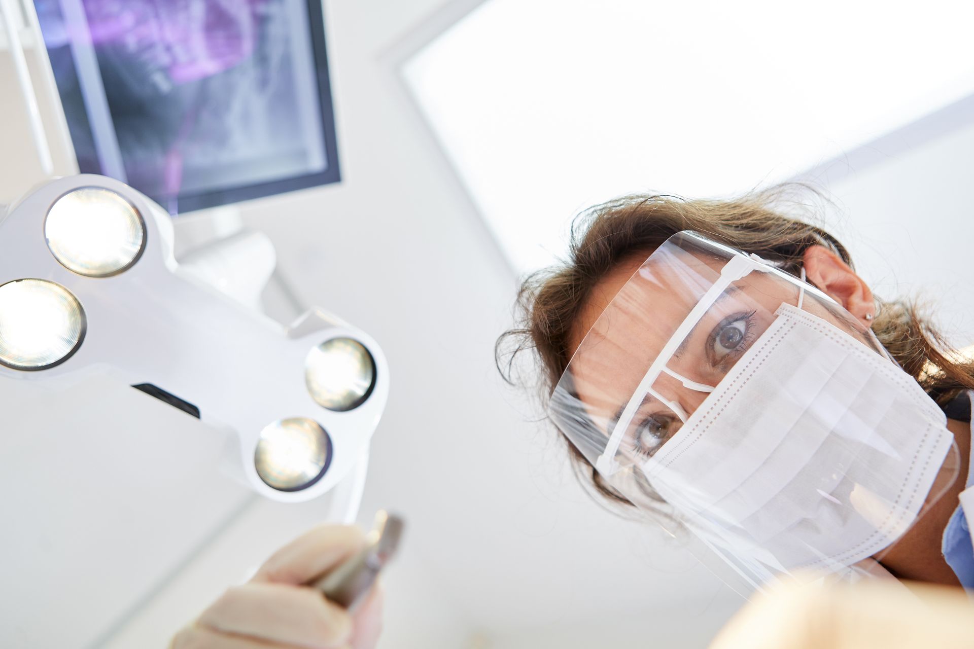 A dentist looking down at the camera with a surgical mask and visor. A dentist looking down at the camera with a surgical mask and visor.