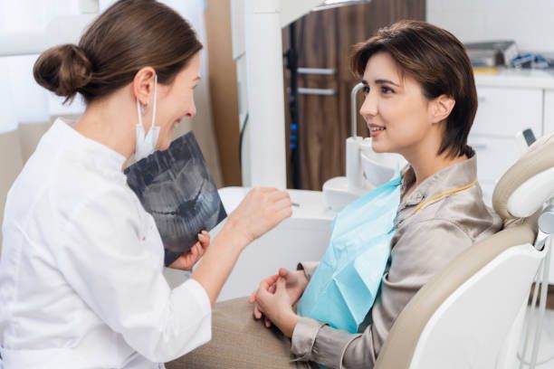 A professional in a white coat shows an X-ray to a patient sitting in a dental chair during a clinical consultation.