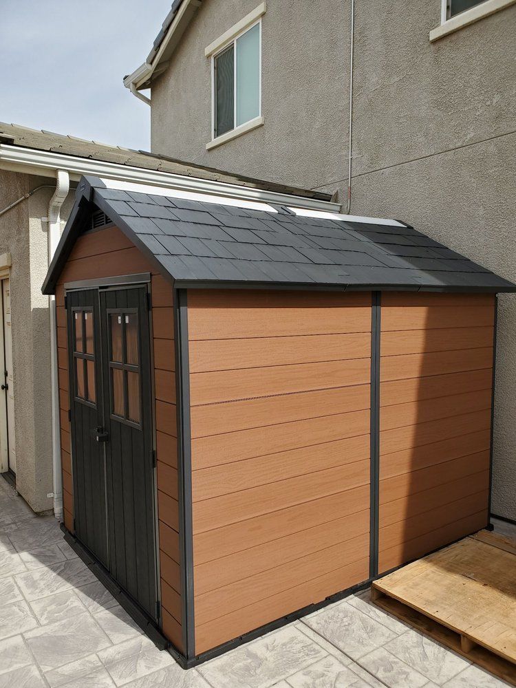 A brown shed with a black roof is sitting next to a house.