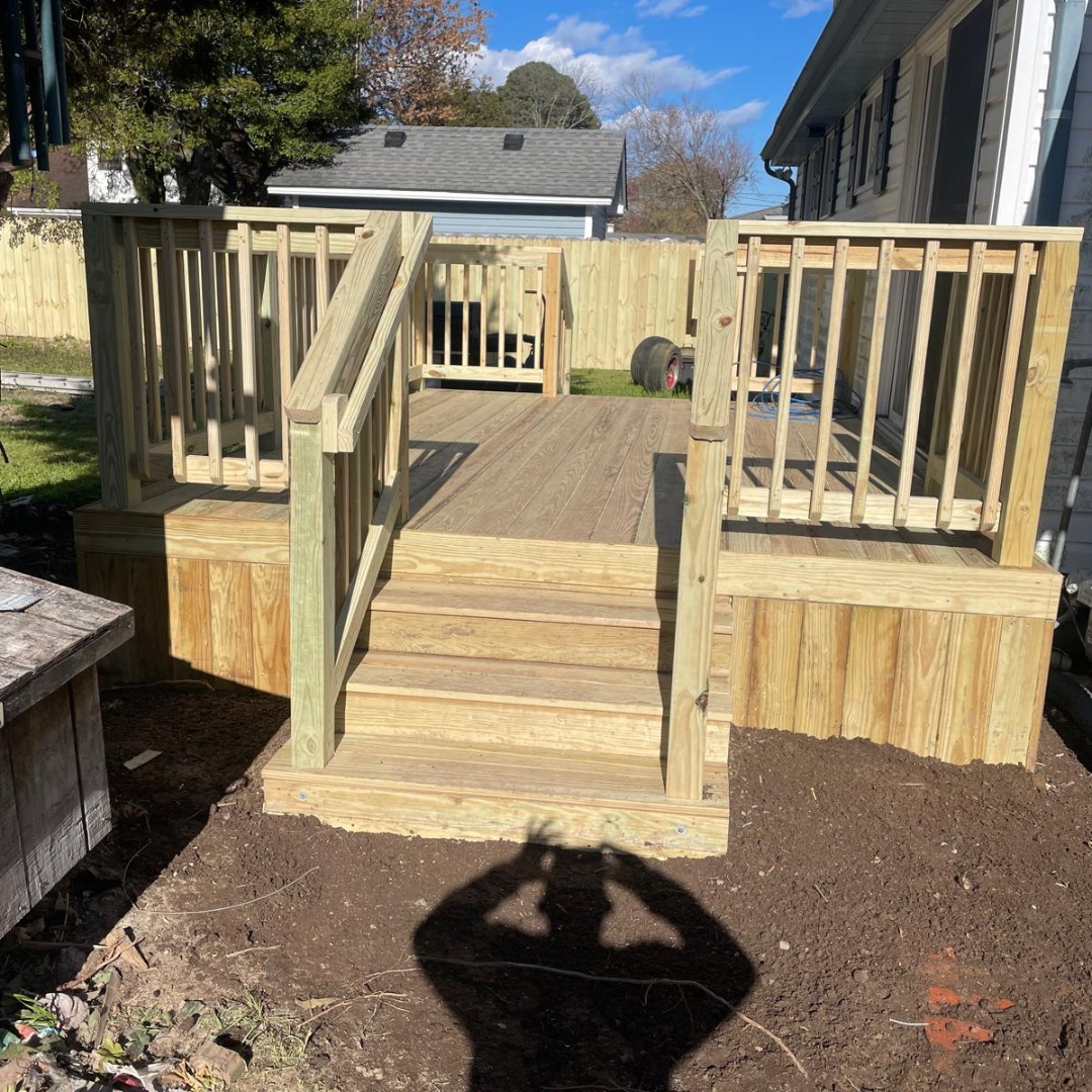 Wooden deck with steps and railings in a yard, with a person's shadow on the ground.