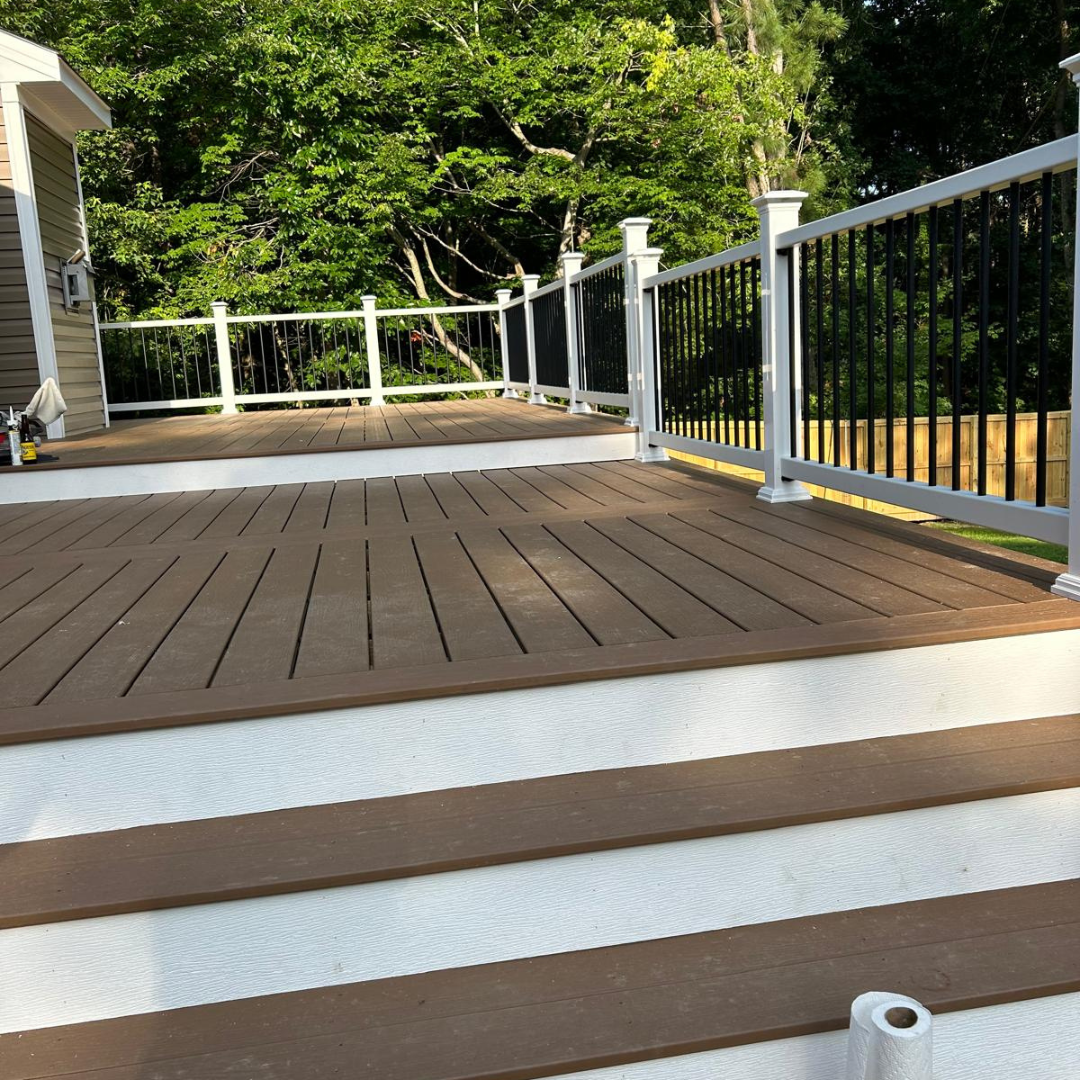 Brown deck with white steps and railings, black metal railing sections, and lush green trees.