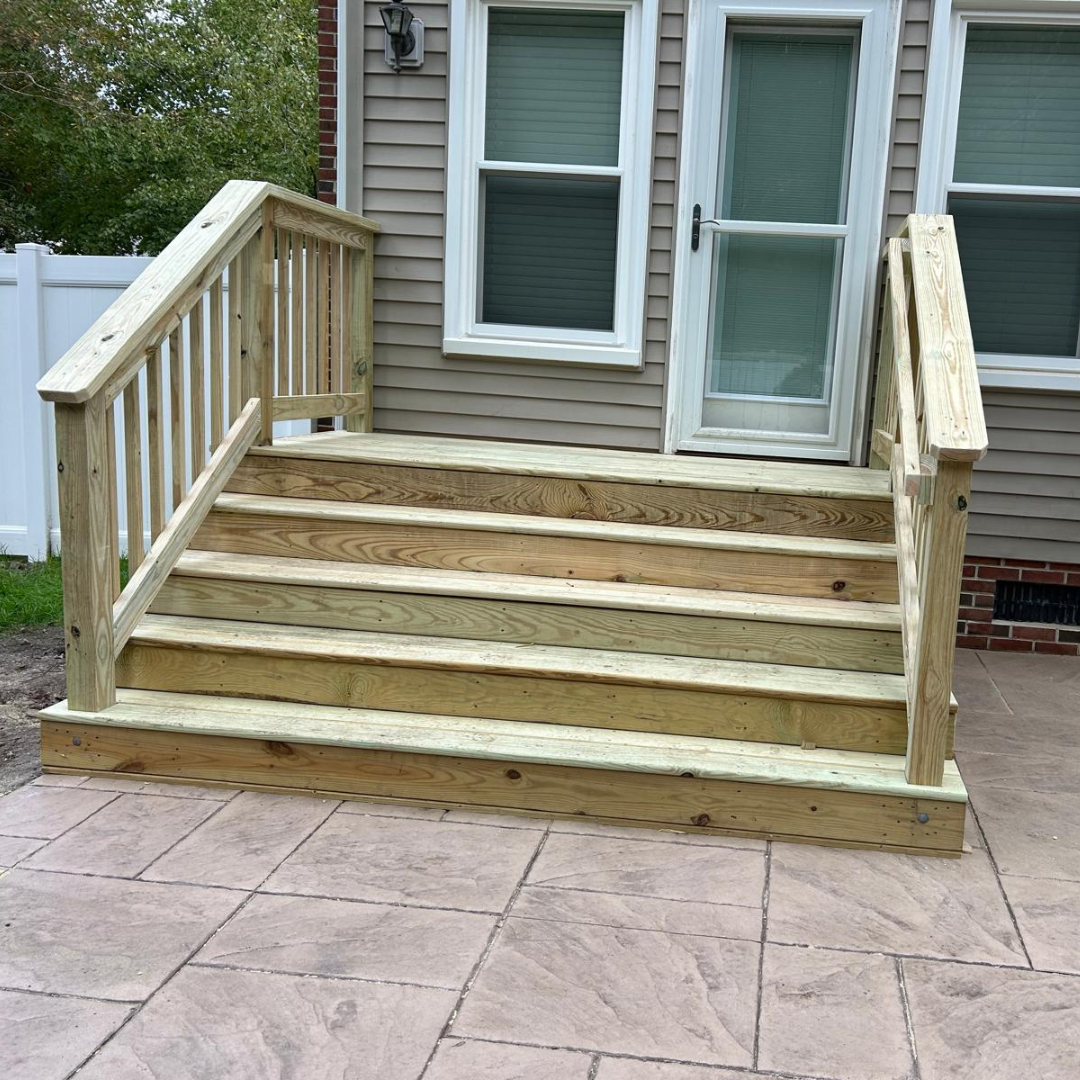 Wooden outdoor steps with railings leading to a doorway. Steps are light brown, set on a stone patio.