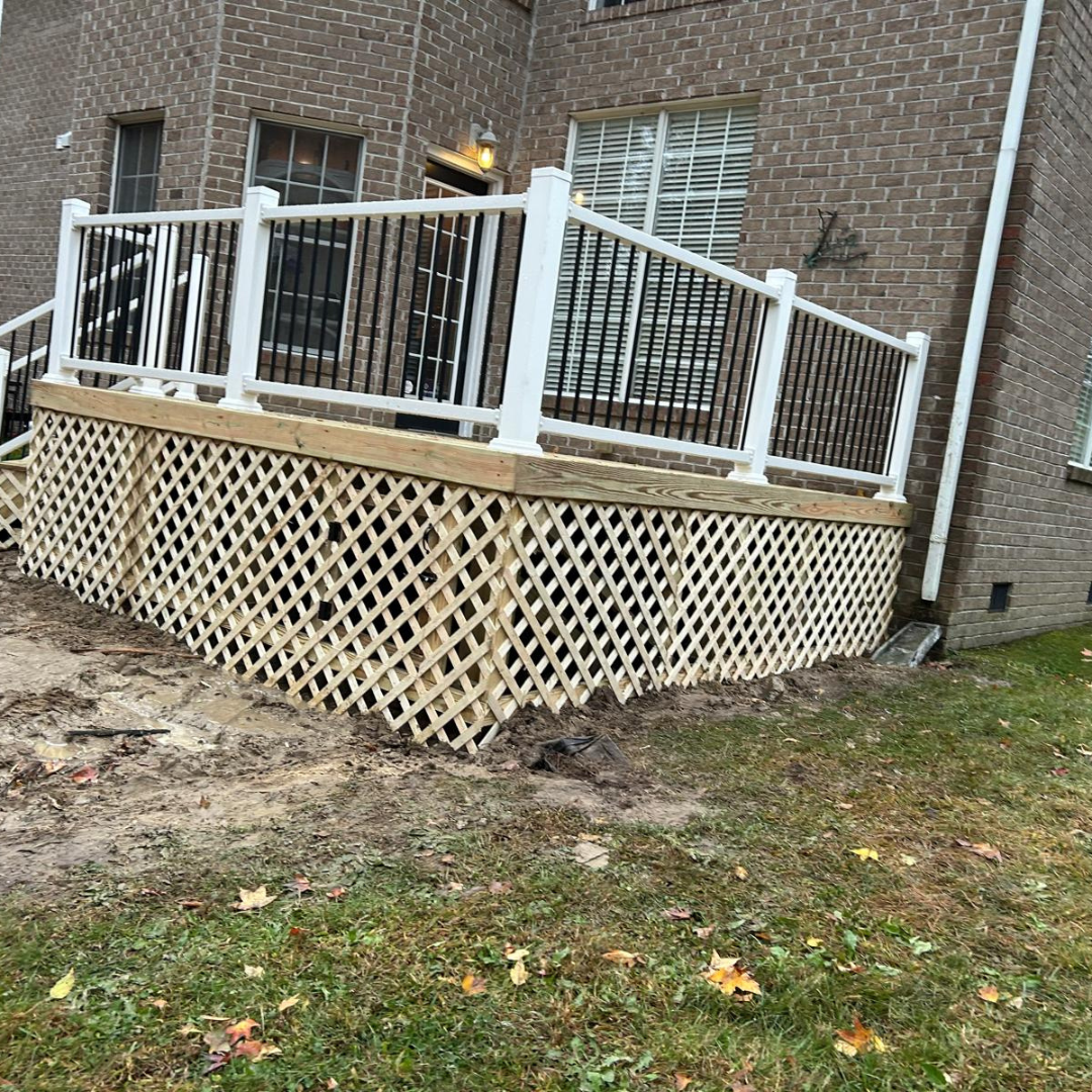 A raised wooden deck with lattice skirting, white railings, and black spindles. Set against a brick house and grassy yard.
