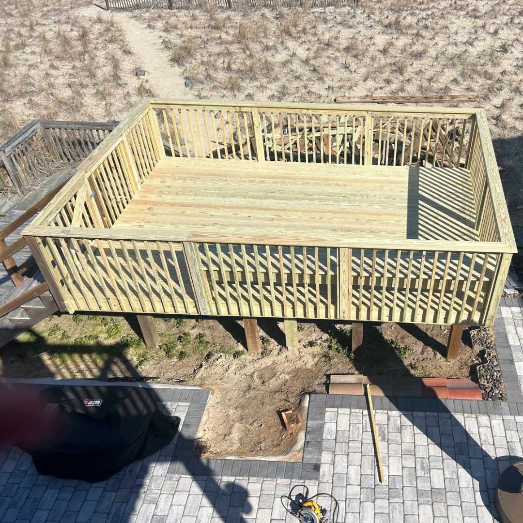 Elevated wooden deck with railing, on posts. Sunlight casts shadows on a stone patio and bare earth below.
