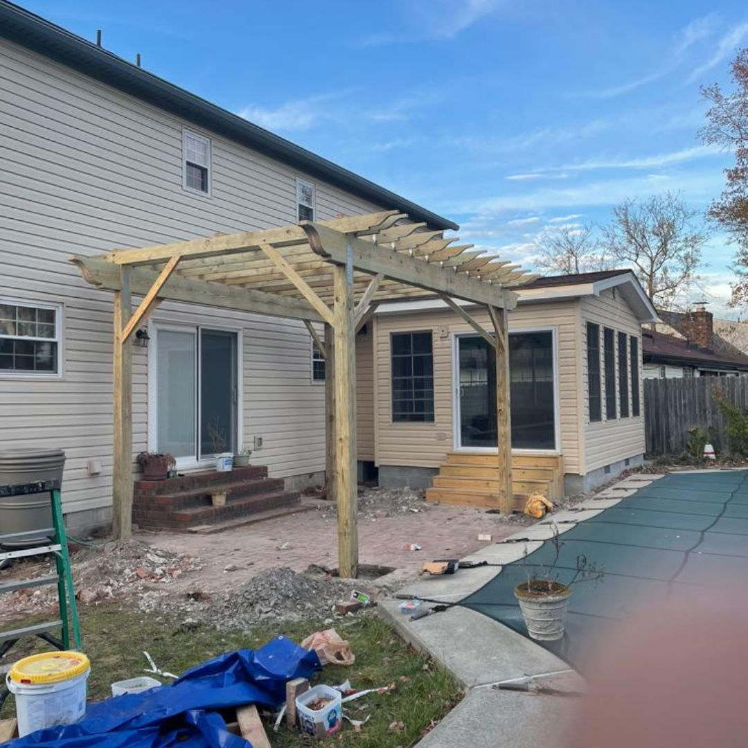 Wooden pergola attached to a house and sunroom. Construction debris and blue tarp on the ground.