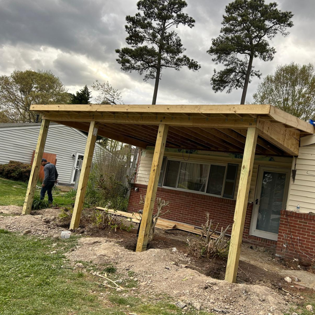Construction of a wooden porch roof on a brick house; person stands near the structure in yard.