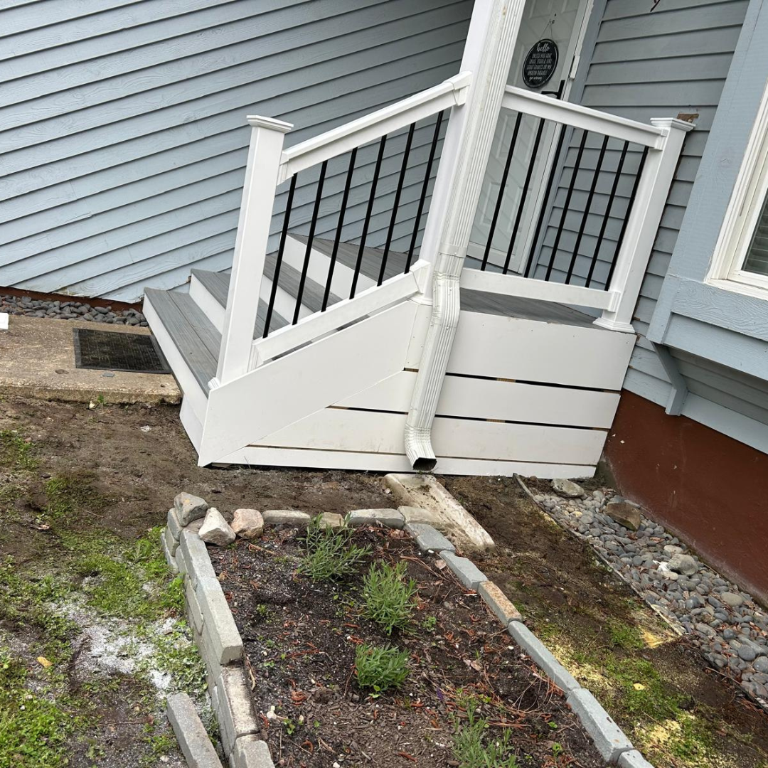 White and gray deck stairs with black railings outside a light blue house, next to a raised garden bed.